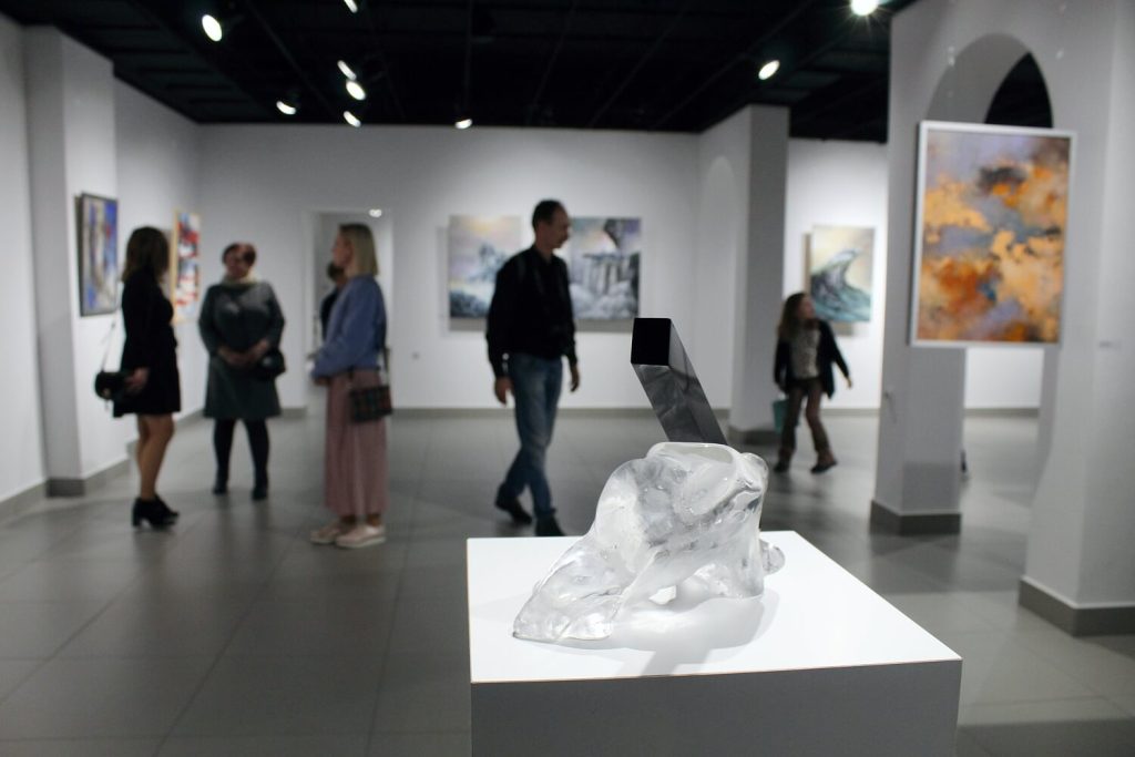 Na pierwszym planie szklana rzeźba w tle zwiedzający wystawę